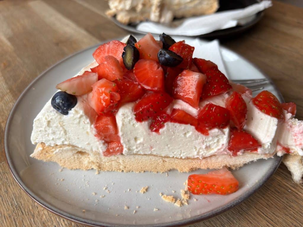 Ein Stück Erdbeerkuchen aus dem Lodz Bistro mit einer dicken Schicht weißer Creme, frischen Erdbeerstücken und Blaubeeren auf einem hellen Baiserboden.