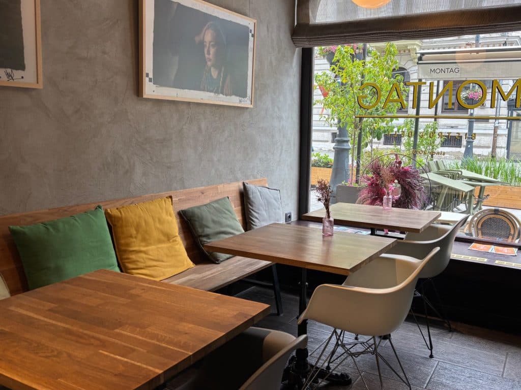 Der modern gestaltete Gastraum des Bistro Montag in Lodz zeigt Holztische mit bunten Kissen auf einer Wandbank sowie weiße Schalensitze vor einem großen Fenster mit Blick nach draußen. An der grauen Wand hängen gerahmte Bilder, die zusammen mit kleinen Blumenvasen auf den Tischen eine gemütliche Atmosphäre schaffen.