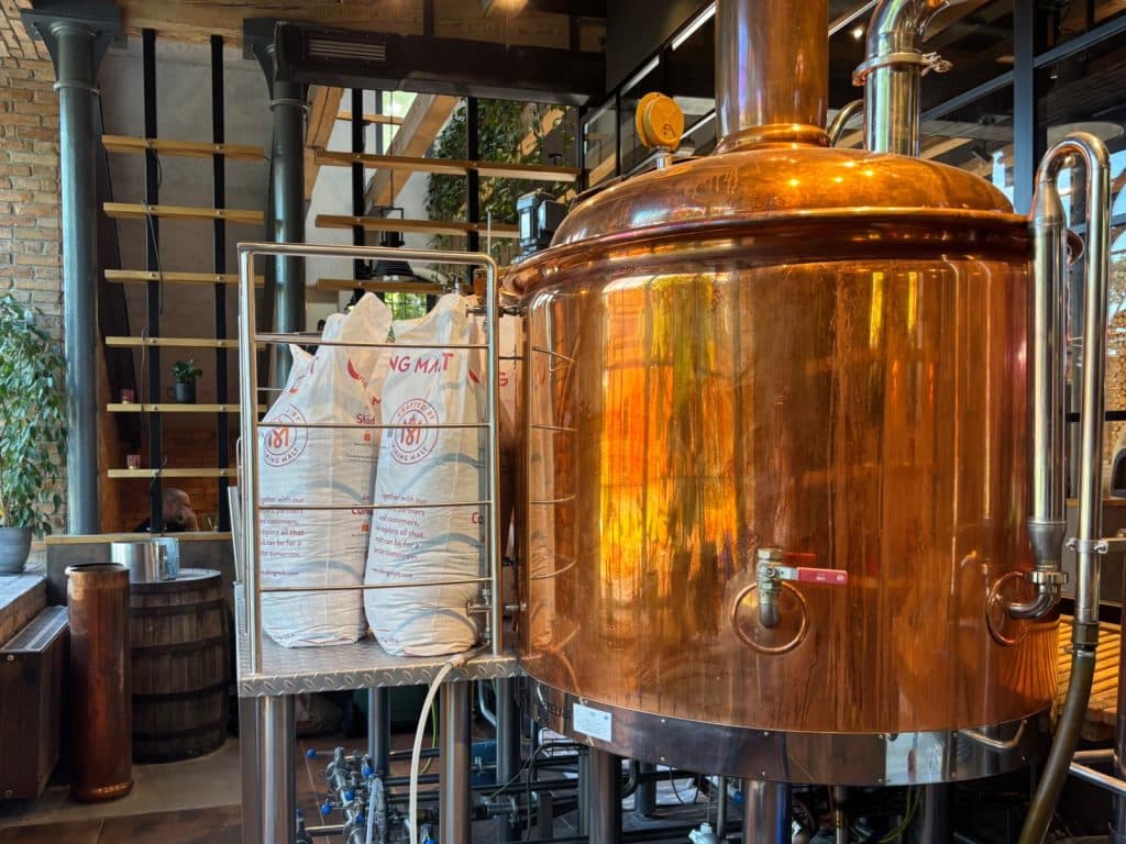 Ein glänzender kupferner Brautank der Brauerei Browar Księży Młyn in Łódź steht neben zwei großen Säcken mit Braumalz auf einer Metallplattform in einem rustikalen Innenraum.
