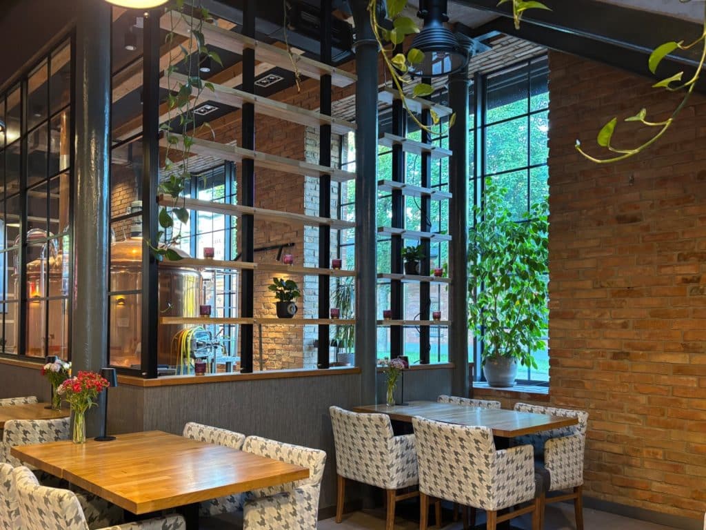 Innenansicht der Brauerei Browar Księży Młyn in Łódź mit rustikalen Backsteinwänden, hölzernen Tischen, gemusterten Sesseln und großen Fenstern, hinter denen Braukessel und grüne Pflanzen sichtbar sind.