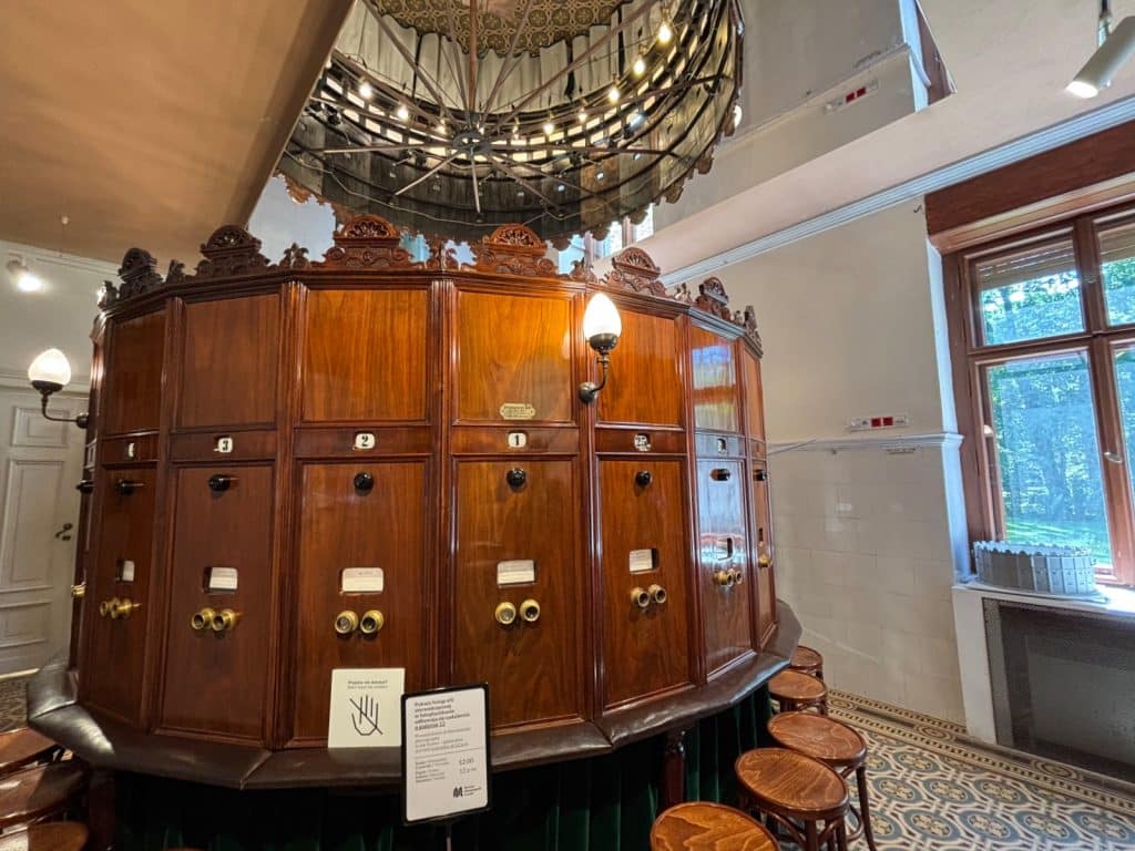 Ein kreisförmiges, hölzernes Kaiserpanorama im Filmmuseum in Łódź mit mehreren nummerierten Sichtstationen, Messingbeschlägen und davor angeordneten Hockern unter einem großen Kronleuchter.