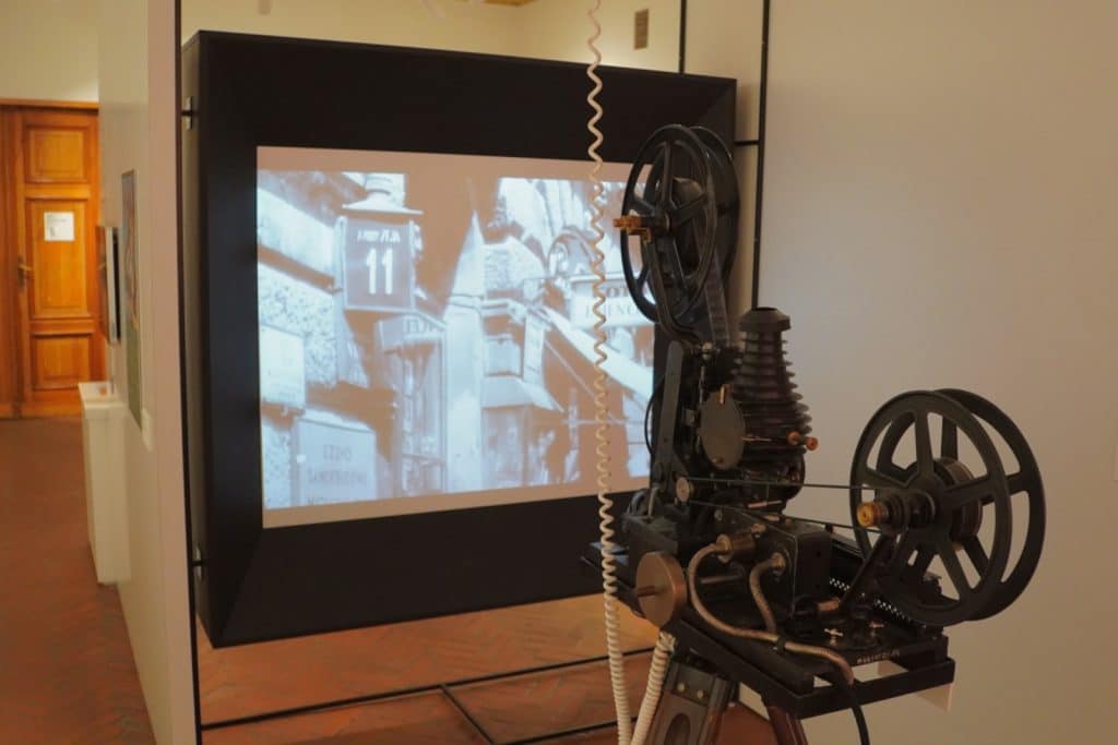 Ein historischer Filmprojektor im Filmmuseum Łódź projiziert einen Schwarz-Weiß-Film auf eine Leinwand, die eine Straßenszene mit der Hausnummer 11 zeigt.