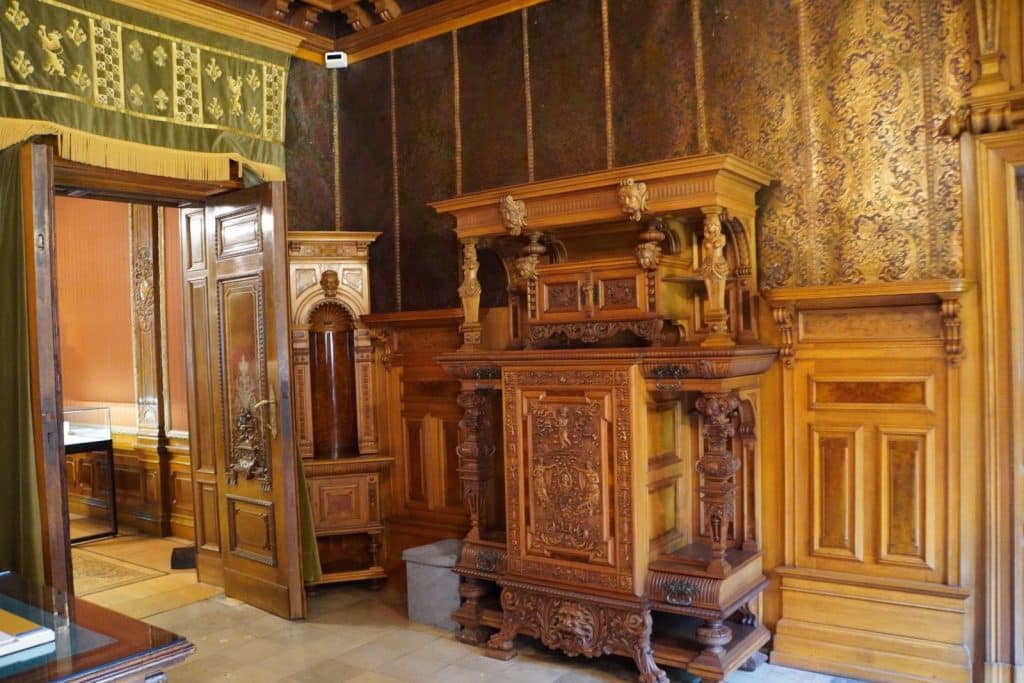 Ein prunkvoll geschnitzter, antiker Holzschrank steht vor einer gemusterten Tapete in einem historischen Raum des Filmmuseums in Łódź, flankiert von einer geöffneten Flügeltür und holzgetäfelten Wänden.