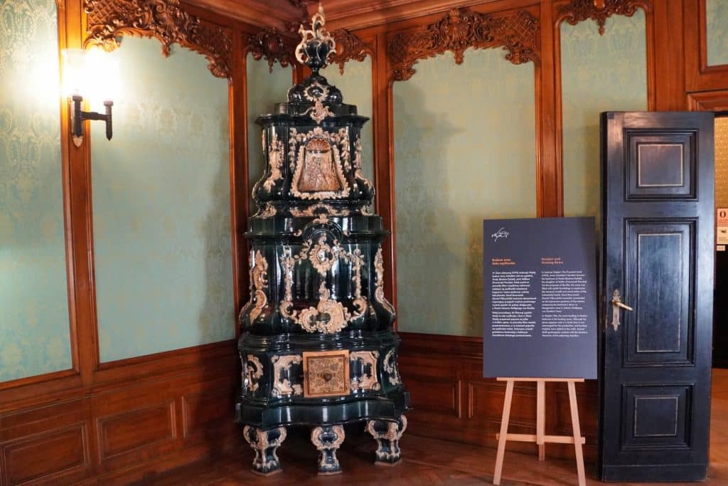 Ein prunkvoller, dunkelgrüner Kachelofen mit hellen Rokoko-Verzierungen steht in einer Zimmerecke des Filmmuseums in Łódź, flankiert von holzgetäfelten Wänden mit hellgrüner Damasttapete und einer Informationstafel.