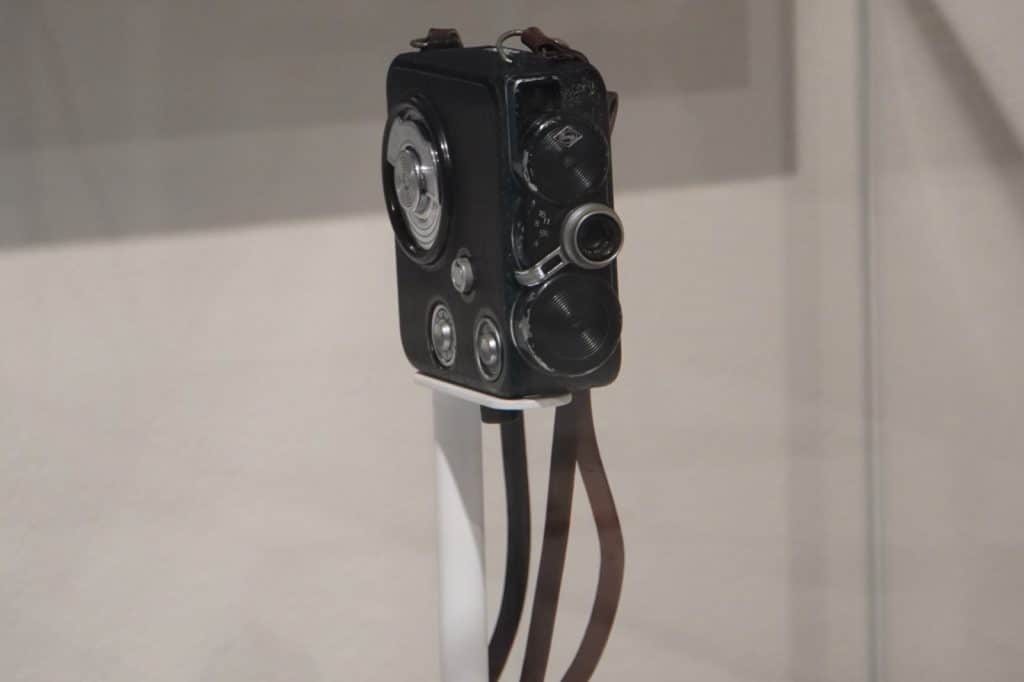 Eine kleine, schwarze Vintage-Filmkamera mit silbernen Bedienelementen und Lederriemen steht auf einer weißen Halterung im Filmmuseum in Łódź.