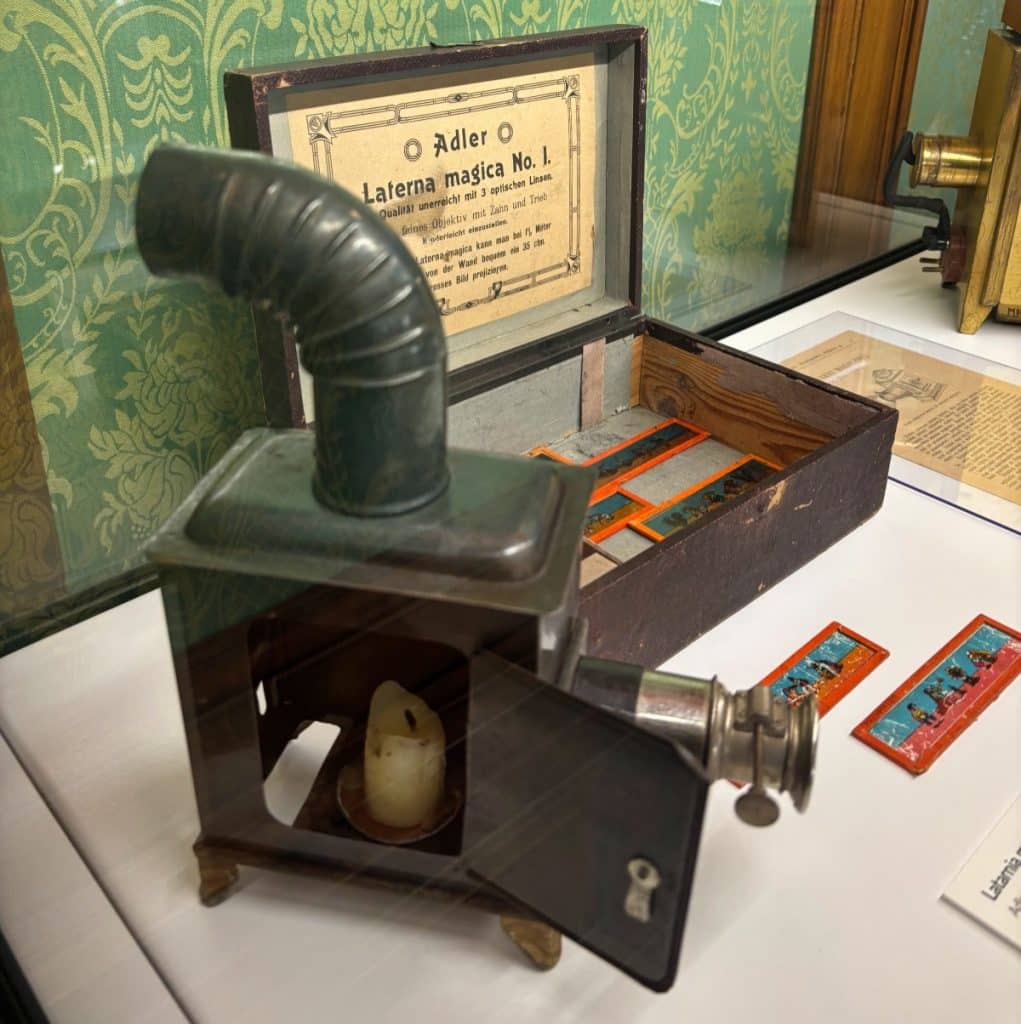 Eine antike Laterna magica aus dem Filmmuseum in Łódź steht mit geöffneter Tür und einer brennenden Kerze im Inneren vor ihrer hölzernen Aufbewahrungsbox, in der bunte Glasdias liegen.
