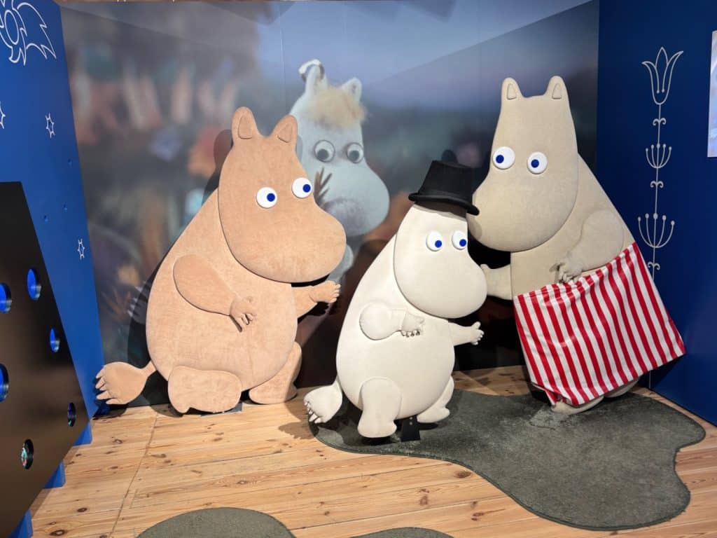 Drei lebensgroße Mumin-Figuren – Muminpapa mit Zylinder, Muminmama mit Schürze und Snorkfräulein – stehen in einer Kulisse im Filmmuseum in Łódź vor einem Hintergrundbild.