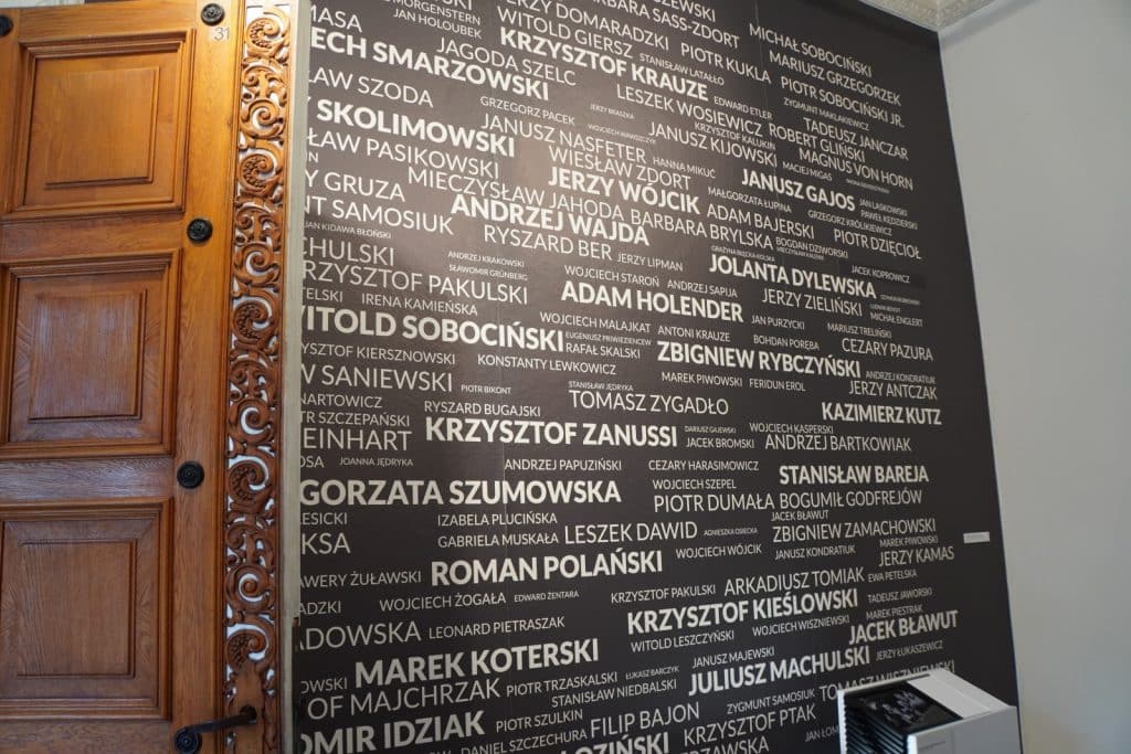 Eine Wand im Filmmuseum in Łódź ist mit zahlreichen Namen polnischer Filmschaffender wie Roman Polański und Andrzej Wajda in weißer Schrift auf schwarzem Grund gestaltet, direkt neben einer kunstvoll geschnitzten Holztür.