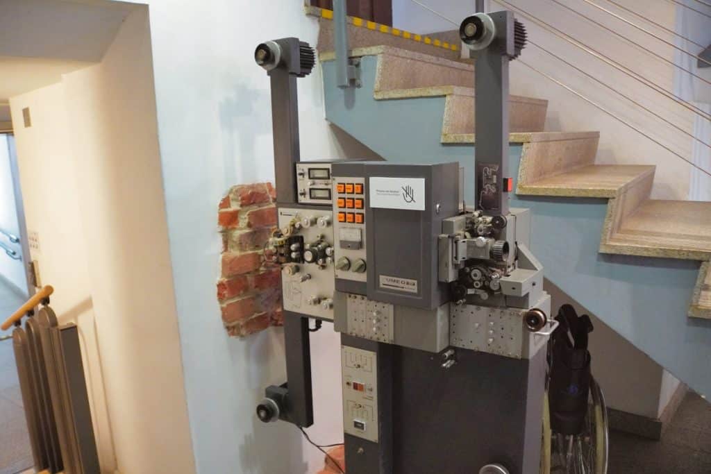 Ein massiver, grauer Filmprojektor mit zahlreichen Knöpfen, Schaltern und Filmspulenarmen steht im Lodz Filmmuseum neben einer Treppe vor einer hellen Wand mit freigelegten Ziegelsteinen.
