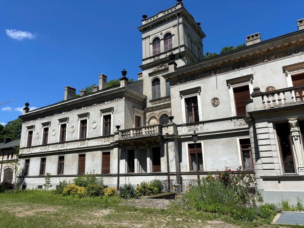 Die Gartenseite des Scheibler-Palastes in Lodz, in dem sich das Filmmuseum befindet, zeigt eine prachtvolle, hellgraue Fassade im Neorenaissancestil mit einem markanten Turm und einem verzierten Balkon vor einem blauen Himmel.