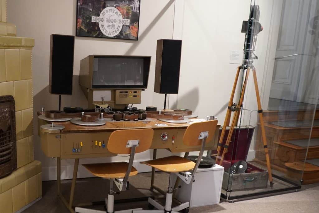 Ein historischer Schneidetisch mit Monitor und Filmspulen im Filmmuseum Łódź, flankiert von zwei Holzstühlen und Lautsprechern sowie einer Filmkamera auf einem Stativ daneben.