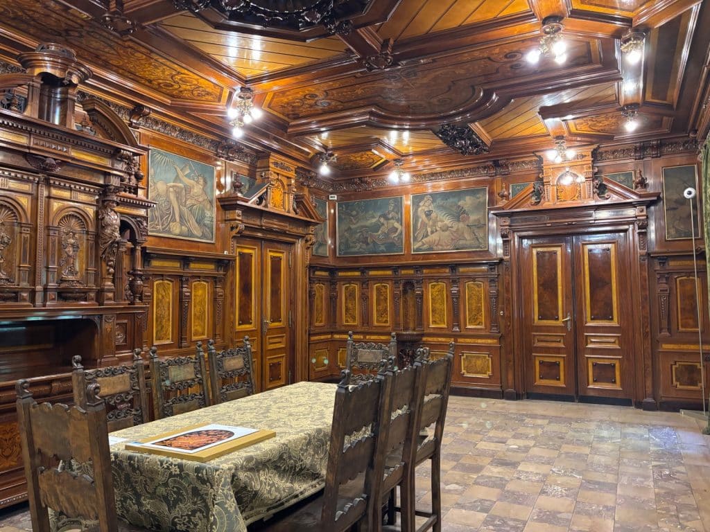 Ein prachtvoller, holzgetäfelter Speisesaal im Filmmuseum in Łódź mit einer kunstvoll geschnitzten Kassettendecke, historischen Wandgemälden und einem langen, mit einer gemusterten Tischdecke gedeckten Esstisch samt verzierten Holzstühlen.