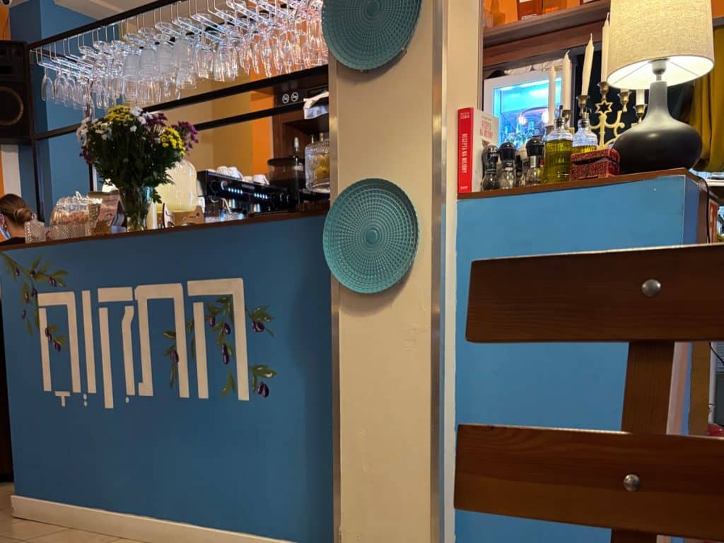 Innenansicht des Restaurants Imber in Lodz mit einer blau gestalteten Theke, die mit hebräischen Schriftzeichen und Olivenzweigen verziert ist, sowie hängenden Weingläsern und dekorativen Tellern an der Wand.