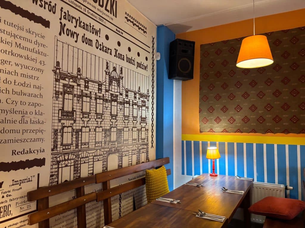Ein gemütlich gedeckter Holztisch im Restaurant Imber in Łódź steht vor einer Wand mit einer großflächigen, historischen Zeitungsseite und farbenfrohen Wandelementen in Blau, Gelb und Orange.