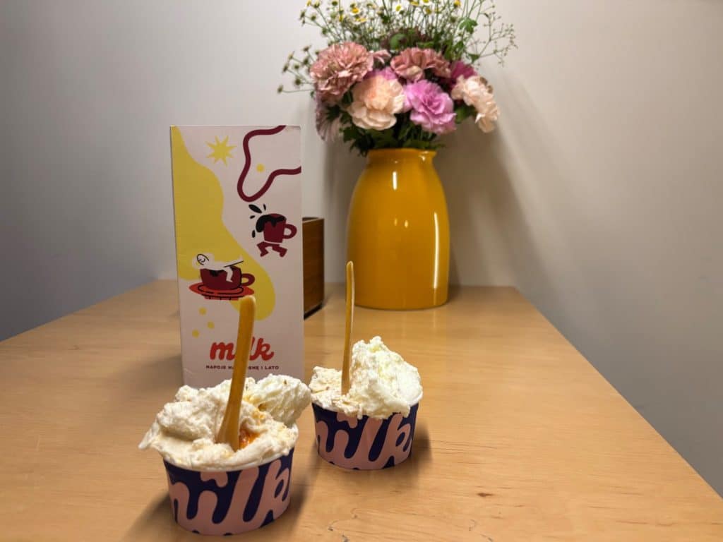 Zwei Becher mit weißem Eis und Holzlöffeln stehen auf einem Holztisch vor einer Speisekarte und einer gelben Blumenvase des Lodz Milk Lodowe Bistro Nawrot.
