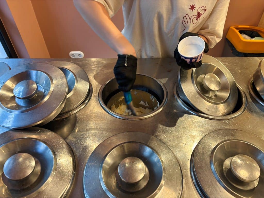 Eine Person mit schwarzen Handschuhen portioniert in der Lodz Milk Lodowe Bistro Nawrot Eis aus einem tiefen Edelstahlbehälter in einen Pappbecher.