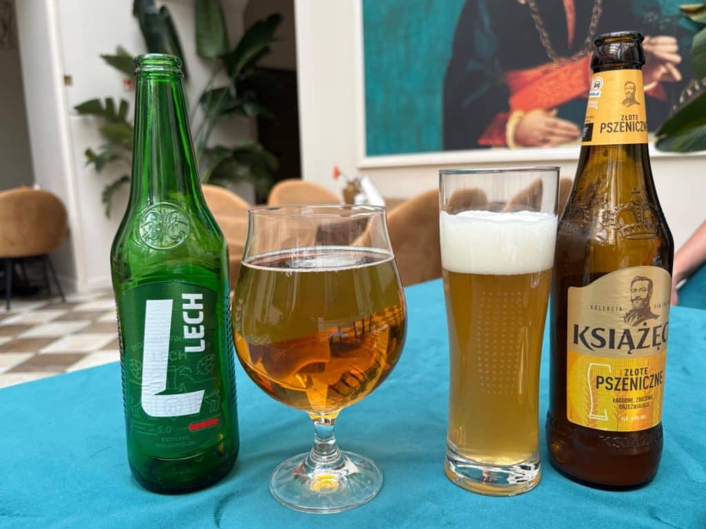 Zwei polnische Biere, ein grünes Lech und ein bernsteinfarbenes Książęce Złote Pszeniczne, stehen mit passend gefüllten Gläsern auf einem blauen Tisch im Restaurant Oranżeria in Łódź.