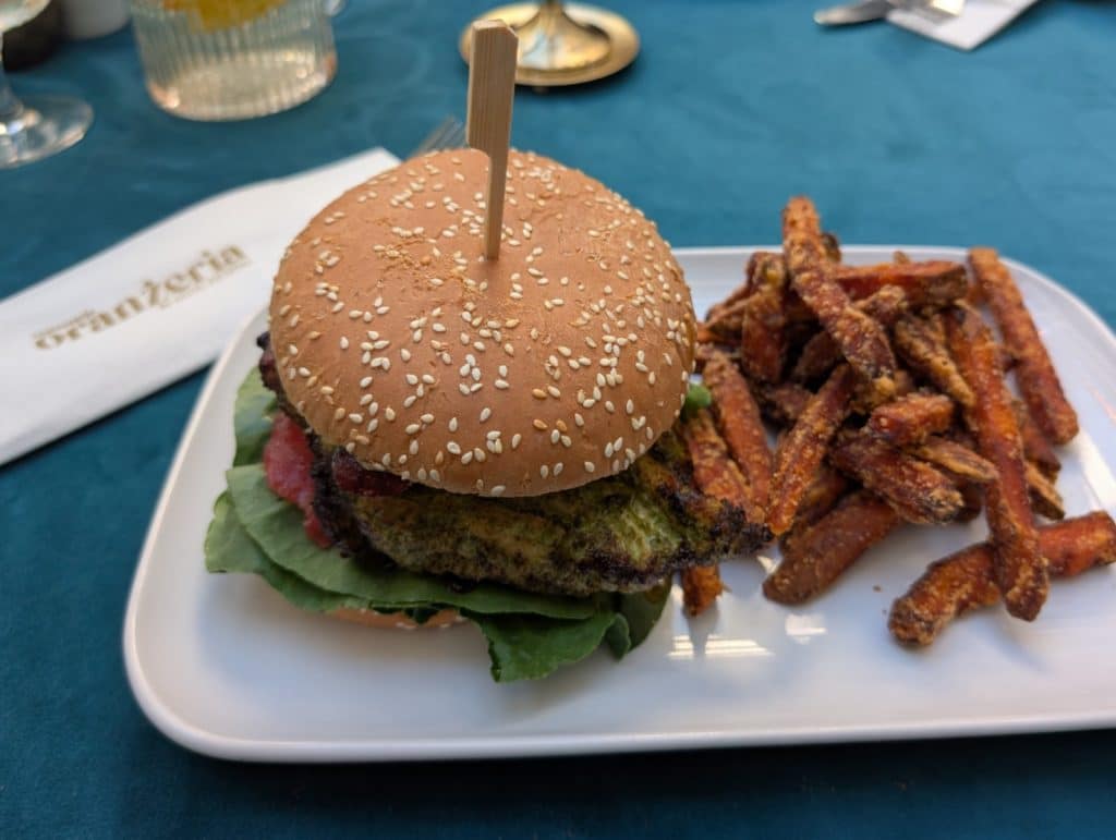 Ein Burger mit Sesambrötchen, grünem Pesto-Belag und Salat wird zusammen mit Süßkartoffel-Pommes auf einem weißen Teller im Restaurant Oranżeria in Lodz serviert.