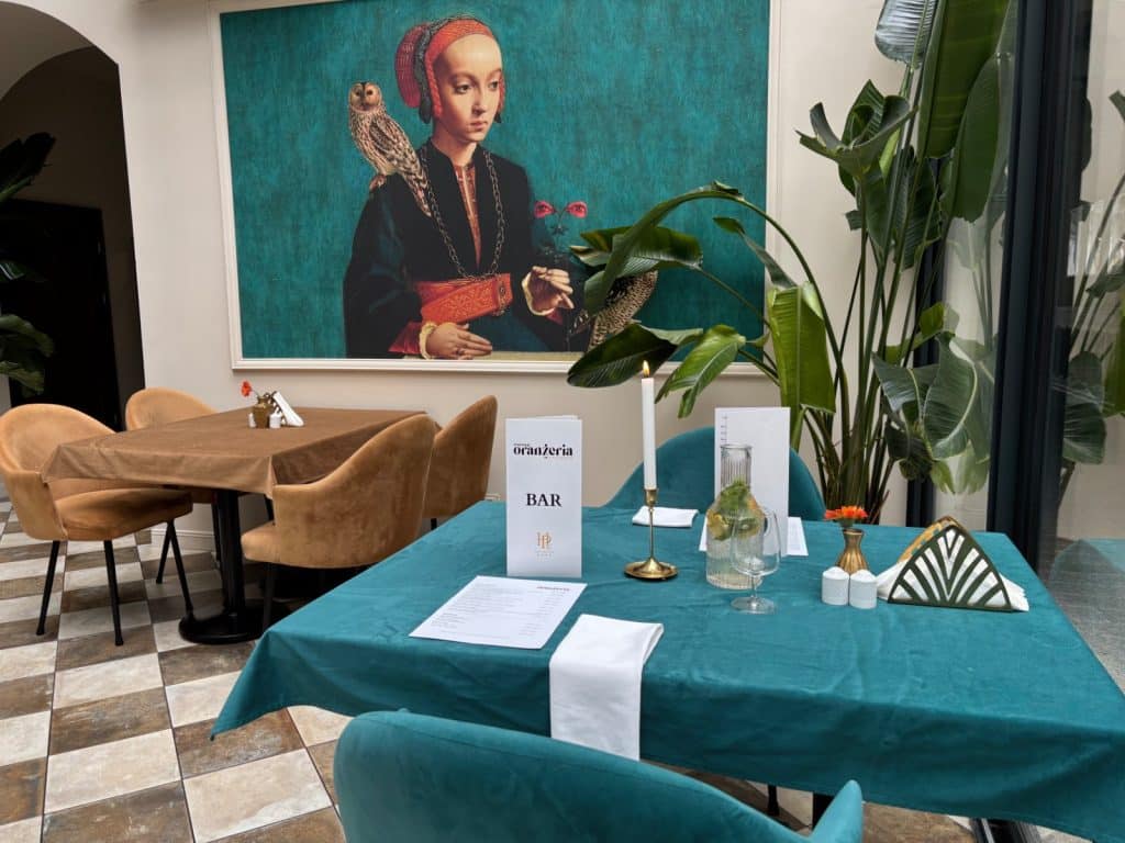 Der Innenraum der Restauracja Oranżeria in Łódź zeigt einen elegant eingedeckten Tisch mit blaugrüner Tischdecke vor einem großformatigen Porträtgemälde einer Frau mit Eule. Samtstühle in Ocker und Blaugrün sowie große Zimmerpflanzen ergänzen das stilvolle Ambiente des Restaurants.