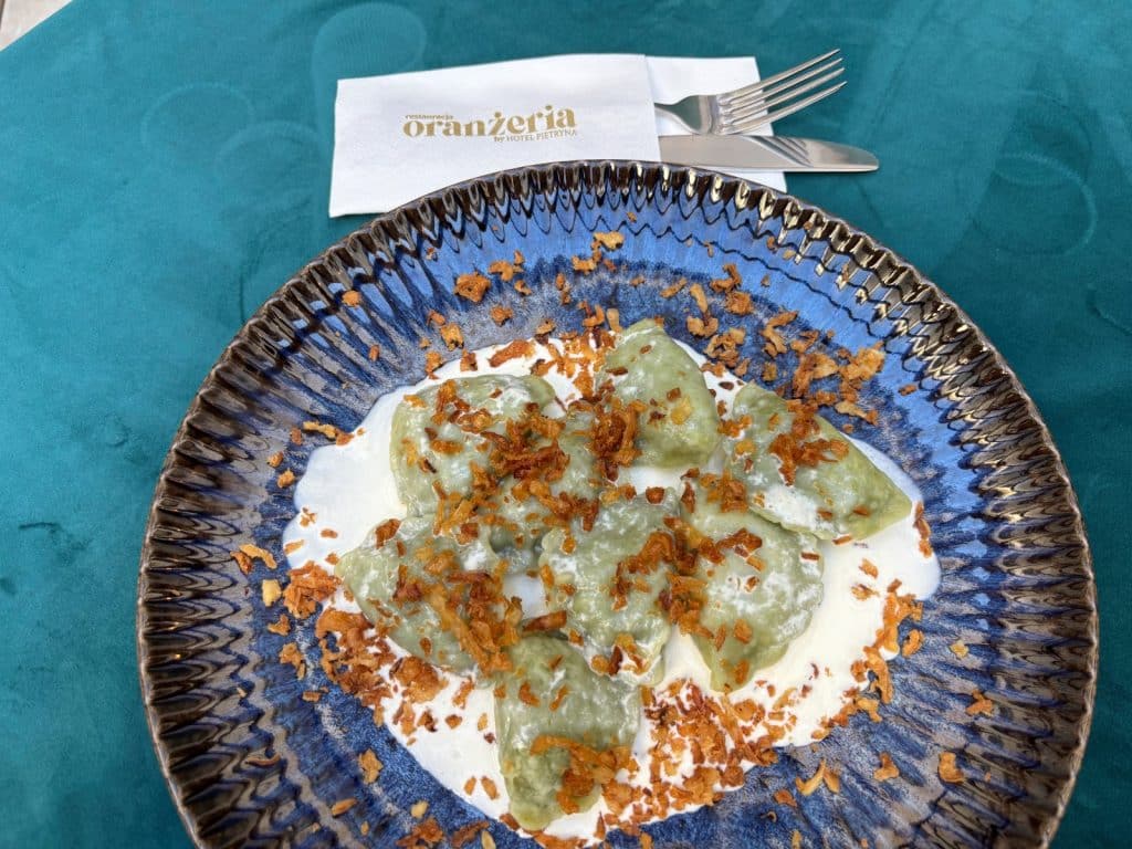 Ein blauer, strukturierter Teller mit grünen Piroggi in einer weißen Sahnesauce, garniert mit Röstzwiebeln, serviert in der Restauracja Oranżeria in Łódź. Neben dem Teller liegen Besteck und eine Serviette mit dem Logo des Restaurants auf einer türkisfarbenen Tischdecke.