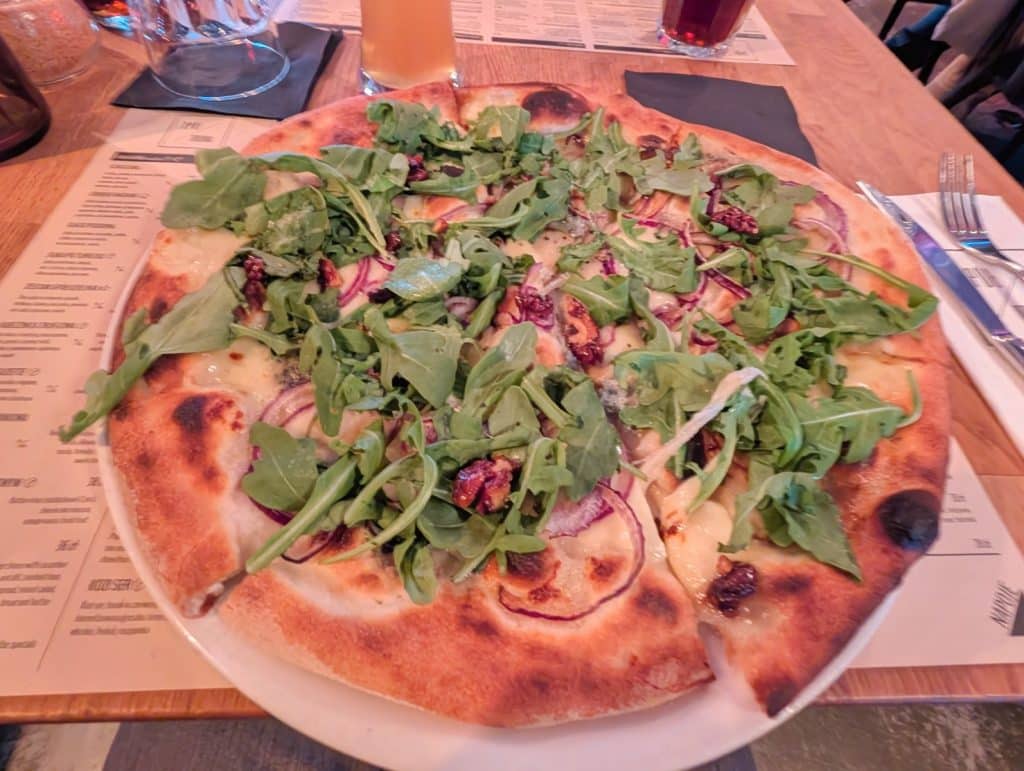 Eine frisch gebackene Pizza im Restaurant Spoldzielnia in Lodz, belegt mit Rucola, roten Zwiebelringen und Walnüssen auf einem hellen Teigboden.