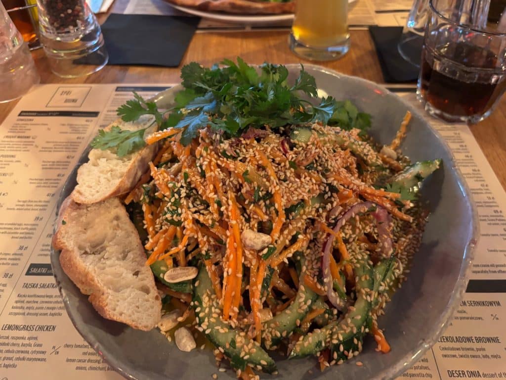 Ein thailändischer Salat im Restaurant Spółdzielnia in Łódź, bestehend aus geraspelten Karotten, Gurkenscheiben und roten Zwiebeln, großzügig mit Sesam bestreut und mit frischem Koriander sowie zwei Scheiben Baguette garniert.