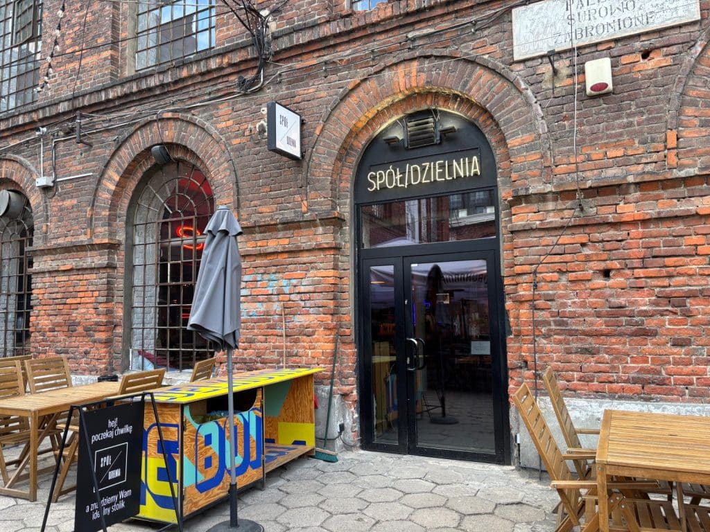 Die Außenansicht des Restaurants Spółdzielnia in Łódź zeigt einen Eingangsbereich mit schwarzer Tür und goldenem Schriftzug in einem rustikalen Backsteingebäude, ergänzt durch hölzerne Außentische und einen bunten Tresen.