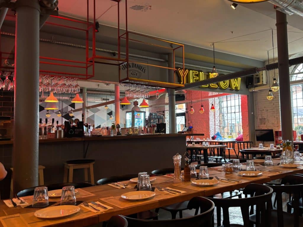 Innenansicht des Restaurants Spółdzielnia in Łódź mit modernem Industrie-Design, langen Holztischen, einer Bar im Hintergrund und auffälligen gelben Neonbuchstaben an der Wand.