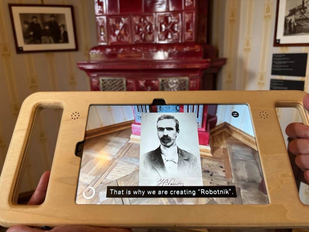Ein Tablet in einer Holzhalterung zeigt eine Augmented-Reality-Anwendung im Museum Bibuła Łódź, bei der ein historisches Porträt des jungen Piłsudski über die reale Museumsumgebung eingeblendet wird, begleitet vom Untertitel „That is why we are creating 'Robotnik'“.