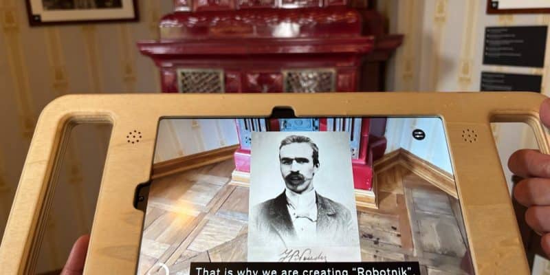 Ein Tablet in einer Holzhalterung zeigt eine Augmented-Reality-Anwendung im Museum Bibuła Łódź, bei der ein historisches Porträt des jungen Piłsudski über die reale Museumsumgebung eingeblendet wird, begleitet vom Untertitel „That is why we are creating 'Robotnik'“.