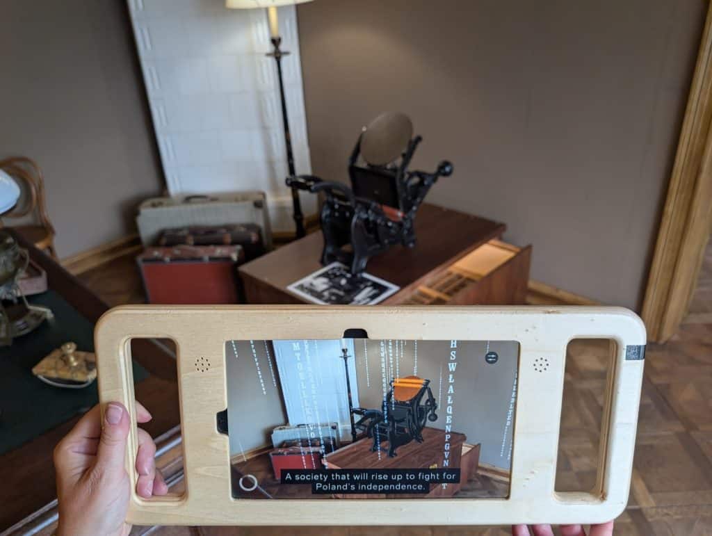 Eine Person hält ein Tablet in einem Holzrahmen vor eine historische Robotnik-Druckmaschine in Łódź, wobei auf dem Display eine Augmented-Reality-Ansicht mit Informationstexten zur polnischen Unabhängigkeit erscheint. Im Hintergrund sind die reale Druckmaschine auf einem Holztisch sowie alte Koffer und eine Stehlampe in einem musealen Raum zu sehen.