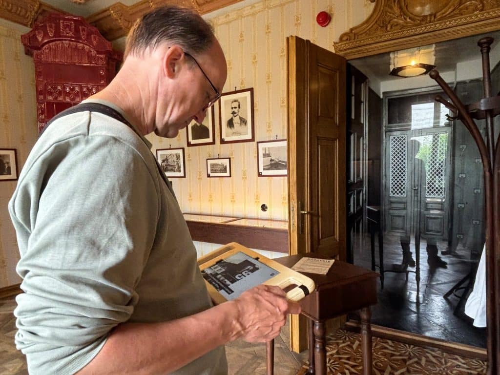 Ein Mann blickt in einem Museum in Lodz auf ein Tablet, das eine historische Ausgabe der Zeitung „Robotnik“ zeigt, während im Hintergrund Porträts an der Wand hängen.
