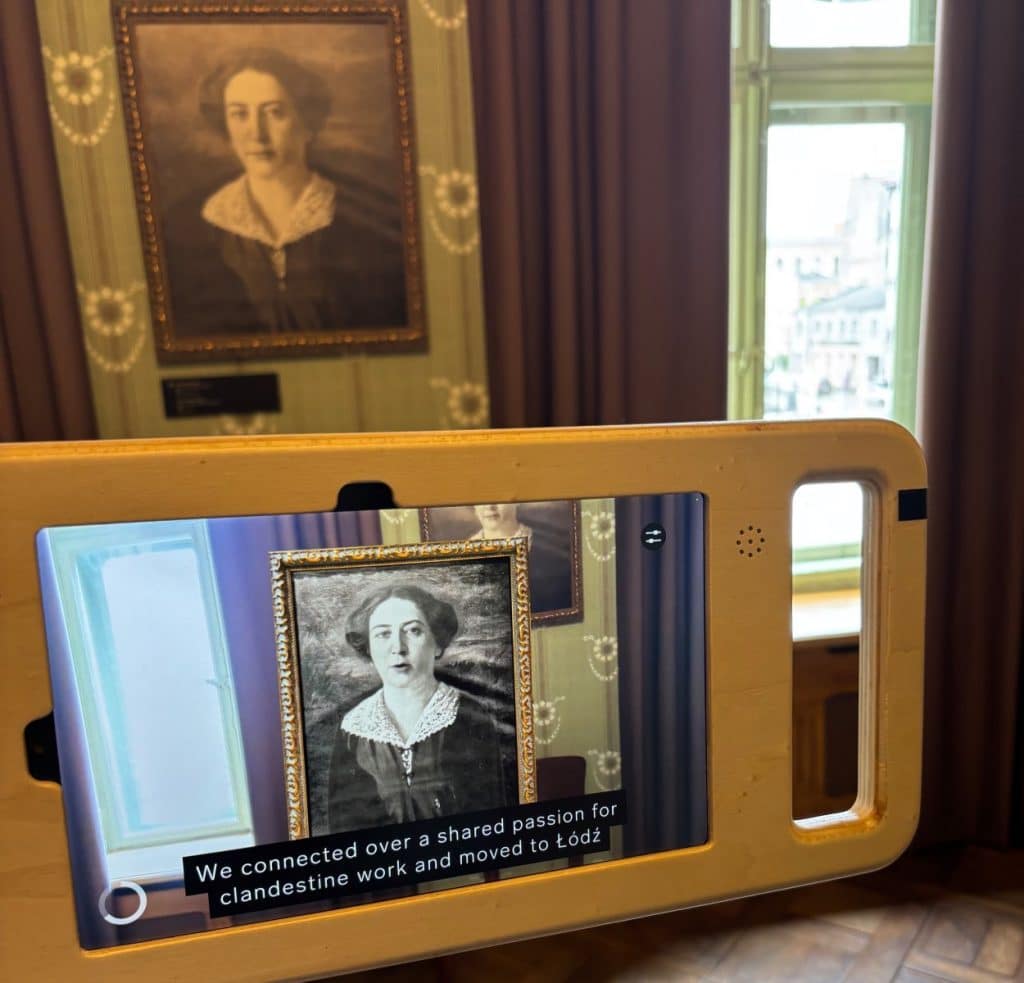 Ein Tablet in einer Holzhalterung zeigt eine Augmented-Reality-Anwendung im Museum Bibuła Łódź, bei der ein historisches Porträt einer Frau zum Leben erweckt wird und Untertitel über ihre clandestine Arbeit erscheinen. Im Hintergrund ist das reale, gerahmte Gemälde an einer gemusterten Wand neben einem Fenster zu sehen.