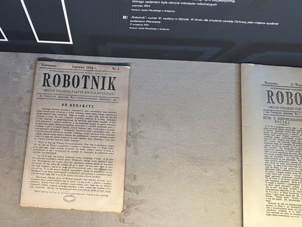Zwei historische Ausgaben der polnischen Zeitung „Robotnik“ aus Łódź liegen nebeneinander in einer Museumsvitrine, wobei die linke Ausgabe vom Juni 1894 datiert ist.
