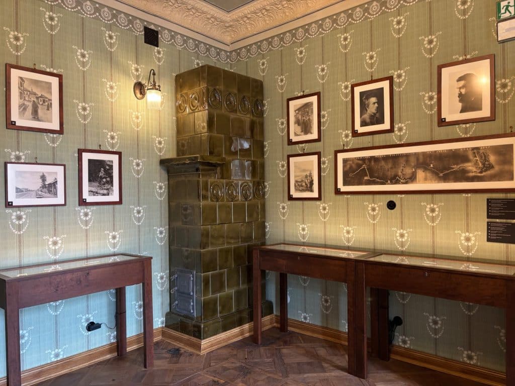 Ein historisches Zimmer im Museum Bibuła Łódź mit gemusterten grünen Tapeten, einem großen grünen Kachelofen in der Ecke und mehreren gerahmten Schwarz-Weiß-Fotografien an den Wänden über hölzernen Vitrinen.