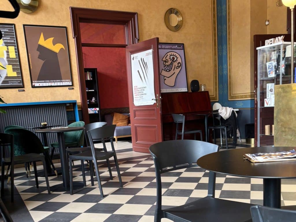 Das gemütliche Innere des Cafés Szykier in Łódź besticht durch einen schwarz-weiß karierten Fliesenboden, dunkle Bistrotische und Wände, die mit künstlerischen Plakaten und einem runden Spiegel geschmückt sind. Eine geöffnete, dunkelrote Holztür führt in einen hinteren Bereich, während ein Klavier und ein Glasregal den künstlerischen Charme des Raumes unterstreichen.