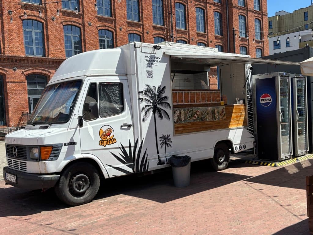 Ein weißer Zapieksa-Foodtruck mit Palmen-Grafiken steht vor einem historischen Backsteingebäude in Lodz, neben einem Pepsi-Getränkekühlschrank.