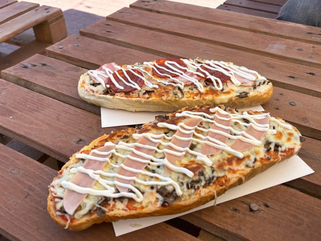 Zwei frisch zubereitete Lodzer Zapiekanki mit geschmolzenem Käse, Pilzen, Schinken und Salami, garniert mit Mayonnaise-Streifen, liegen auf einem rustikalen Holztisch.