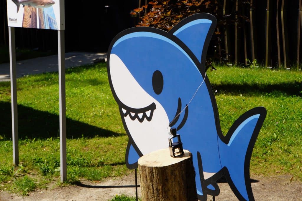 Eine Stempelstation im Orientarium Zoo Lodz mit einem großen, blauen Comic-Hai-Aufsteller und einem auf einem Baumstumpf montierten Metallstempel.