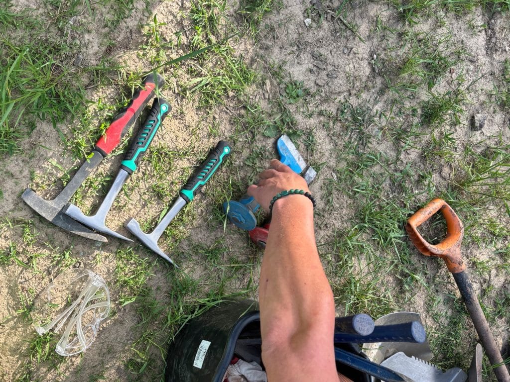 Eine Person greift nach verschiedenen Geologen- und Achat-Hämmern, die zusammen mit einer Schutzbrille und einer Schaufel für die Suche in Niederschlesien auf dem Boden bereitliegen.