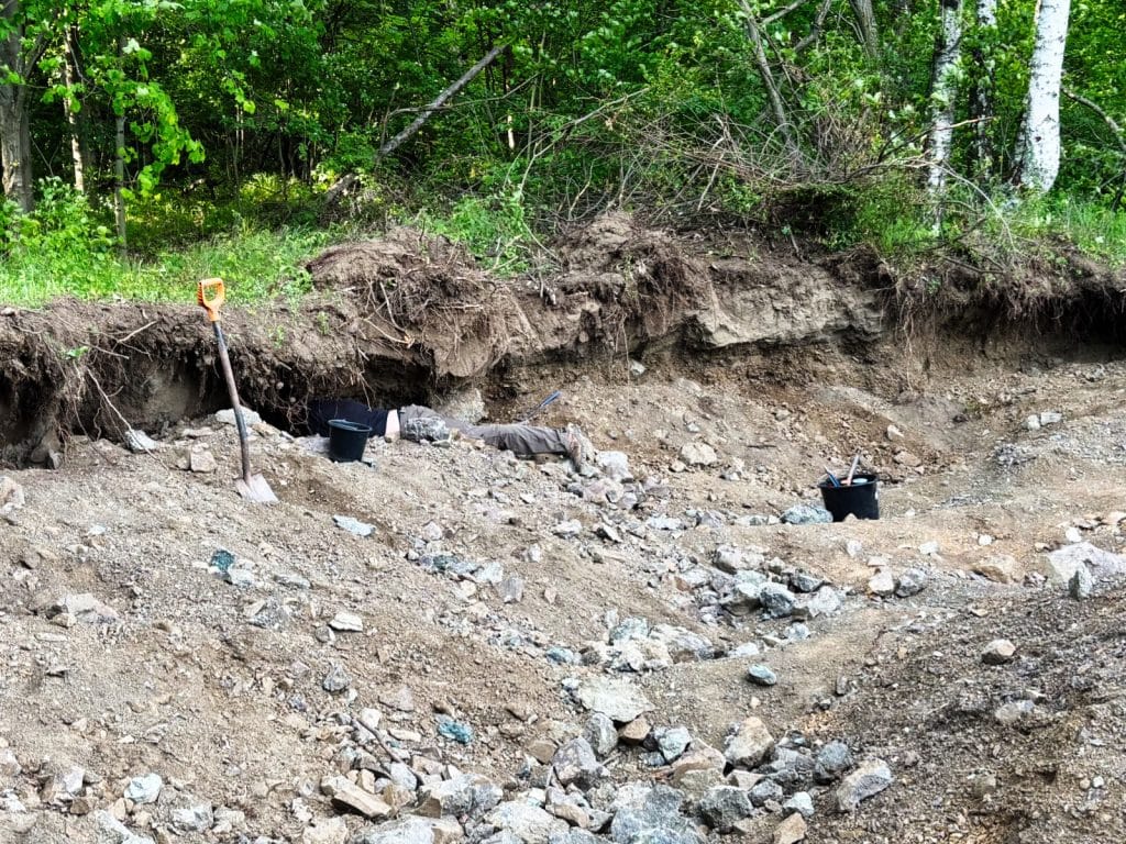 Eine Person liegt bäuchlings in einer Ausgrabungsstätte in Niederschlesien und gräbt unter einem Erdüberhang nach Achaten, umgeben von Schaufeln, Eimern und losem Gestein.