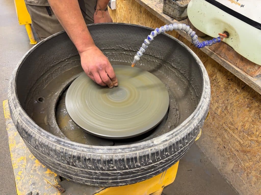 Eine Person schleift in einer Werkstatt einen Achat aus Niederschlesien auf einer rotierenden Schleifscheibe, die in einem alten Autoreifen montiert ist, während Wasser aus einem flexiblen Schlauch zur Kühlung auf den Stein geleitet wird.