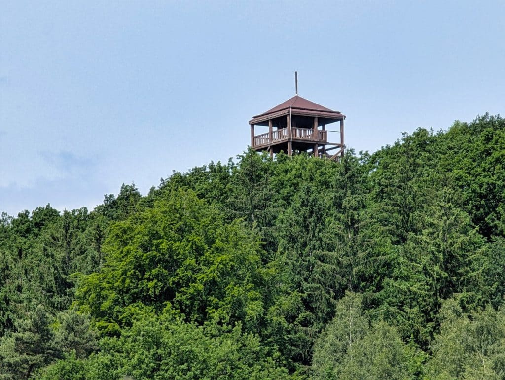 Ein hölzerner Aussichtsturm mit quadratischem Grundriss und spitzem Dach ragt auf einer Radtour in Niederschlesien über die dichten Wipfel eines bewaldeten Hügels.