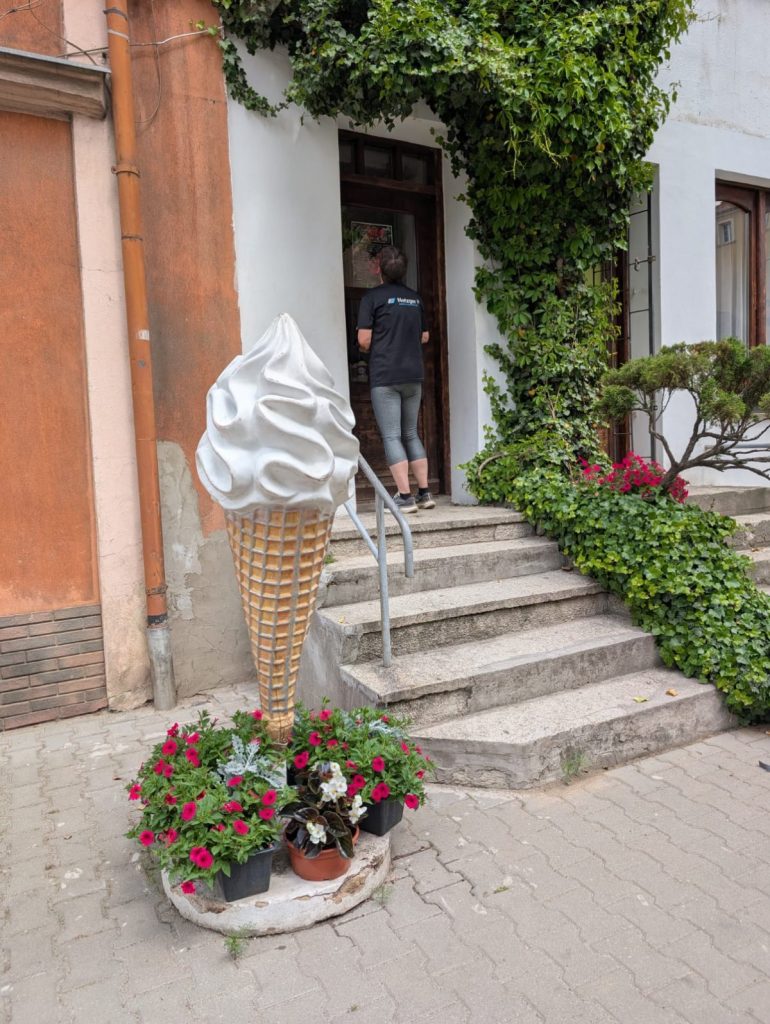 Vor einem Eisladen in Niederschlesien steht eine große Eistüten-Skulptur mit weißem Softeis in einem Blumenbeet neben einer Treppe, die zu einem mit Efeu bewachsenen Eingang führt. Eine Person in Sportkleidung steht vor der Tür des Gebäudes.