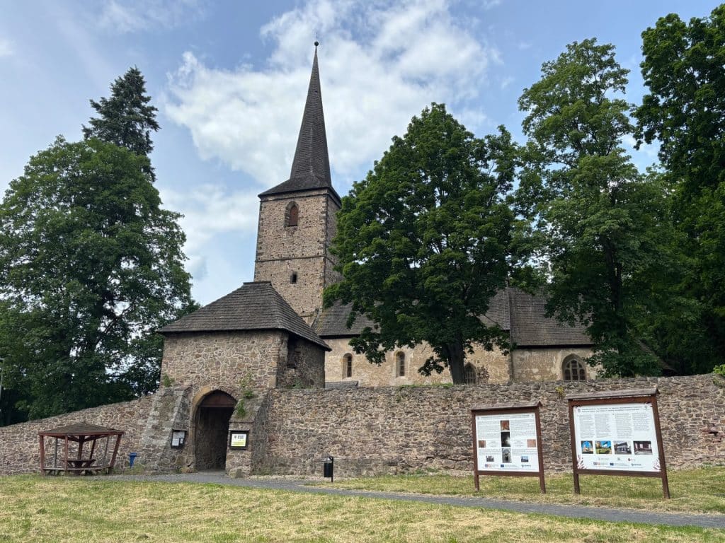 Eine mittelalterliche Steinkirche mit hohem, spitzem Turm in Niederschlesien, umgeben von einer massiven Steinmauer mit Torhaus und Informationstafeln an einem Radweg.