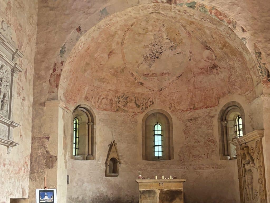 Innenansicht einer historischen Kirche in Niederschlesien mit verblassten mittelalterlichen Wandmalereien in der Apsis, drei Rundbogenfenstern und einem schlichten Steinaltar.