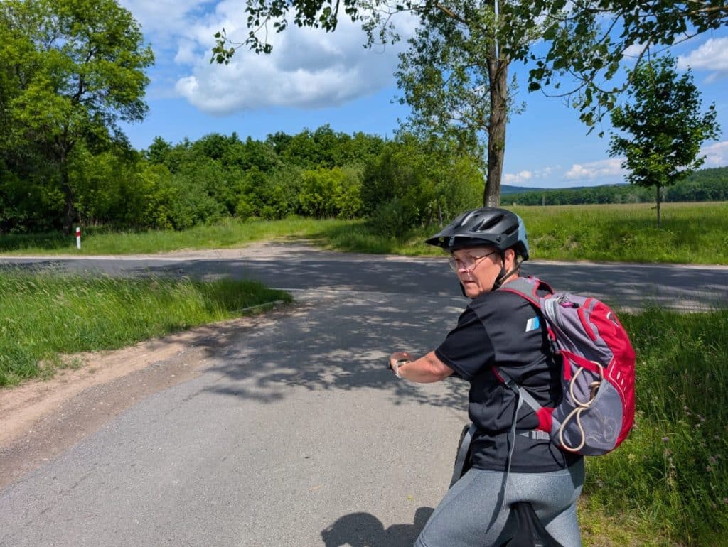 Eine Radfahrerin mit Helm und rotem Rucksack blickt an einer sonnigen Wegkreuzung in Niederschlesien über ihre Schulter zurück.