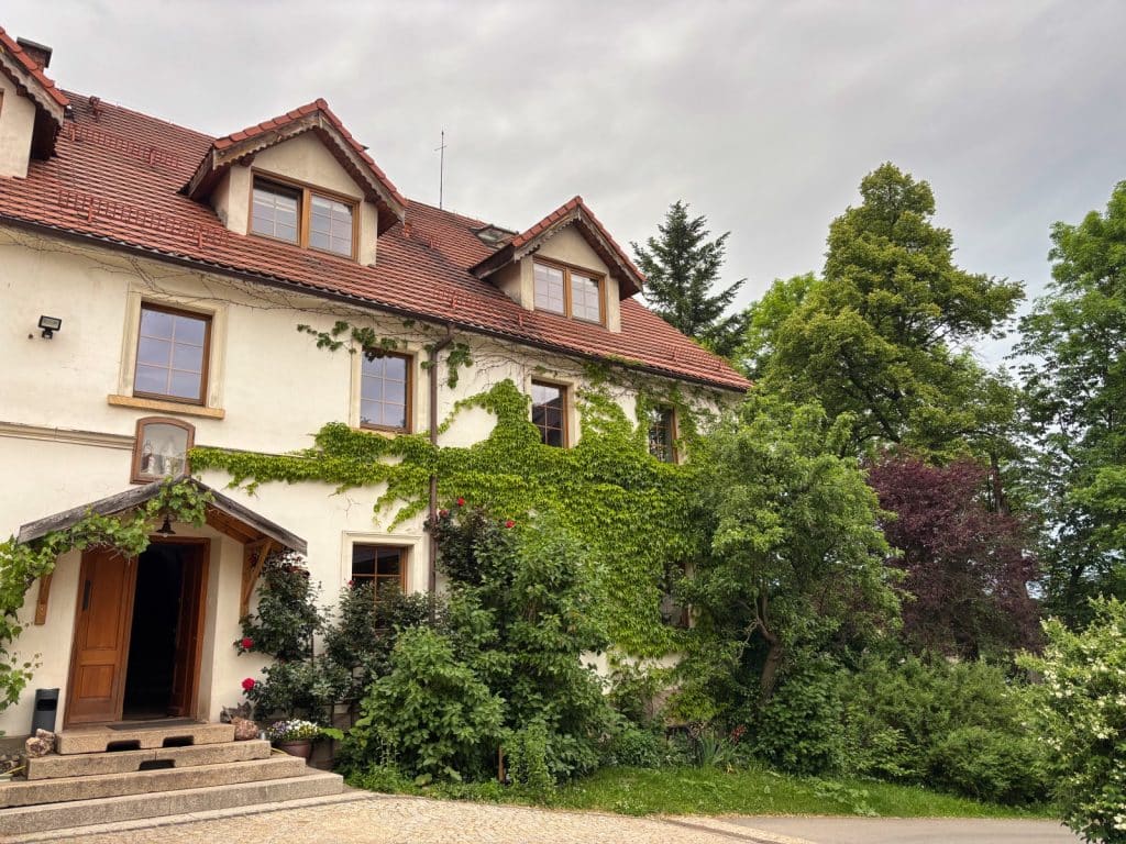 Die Villa Greta in Niederschlesien präsentiert sich als ein helles, zweistöckiges Gebäude mit rotem Ziegeldach, dessen Fassade teilweise von dichtem, grünem Efeu bewachsen ist. Vor dem Eingang mit einer schweren Holztür und Steinstufen erstreckt sich ein üppiger Garten mit verschiedenen Sträuchern, Blumen und hohen Bäumen unter einem bewölkten Himmel.
