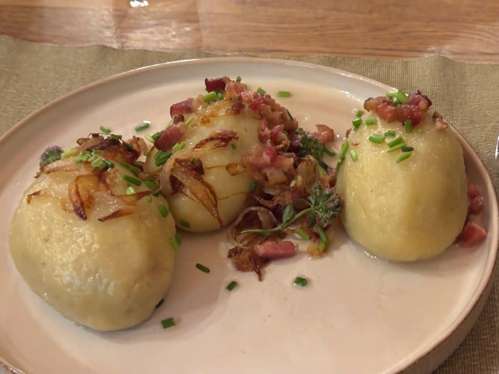 Drei traditionelle niederschlesische Kartoffelklöße, bekannt als „Bomben“, angerichtet auf einem hellen Teller in der Villa Greta und garniert mit gebratenen Zwiebeln, Speckwürfeln sowie frischem Schnittlauch.
