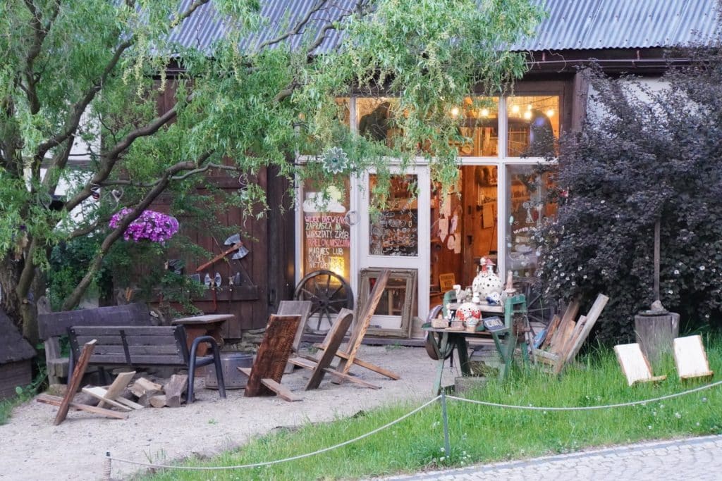 Die Galerie der Villa Greta in Niederschlesien präsentiert sich in einem rustikalen Außenbereich mit Holzstühlen, Bänken und verschiedenen Kunsthandwerksobjekten vor einem charmanten Gebäude mit großen Glasfenstern.